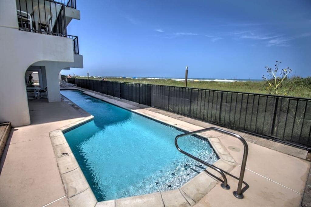 A gated swimming pool is visible adjacent to the beach, featuring a clear blue surface that reflects the sky. A short path leads from the pool to the sandy shoreline, while a black fence surrounds the area for privacy. Sunny skies are seen above.