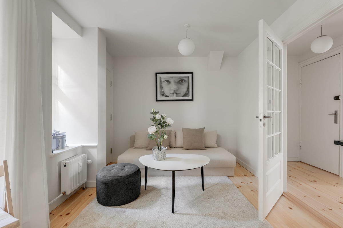 A light and airy living space features a low sofa with neutral cushions, complemented by a round coffee table. A soft area rug lies beneath, and natural light floods in through large windows, highlighting the minimalist decor. A plant adds a touch of greenery to the arrangement.