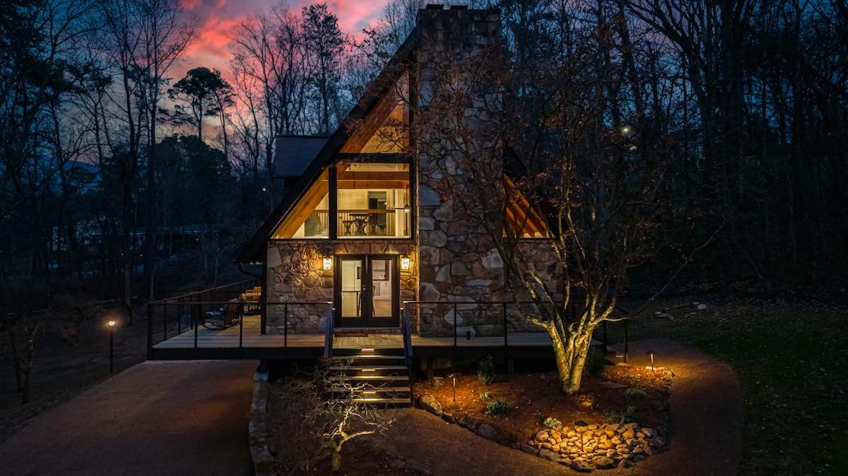 A modern A-Frame cabin is showcased at dusk, featuring a large stone facade and illuminated entrance steps. Soft outdoor lighting highlights the landscaped area, while trees surround the property, creating a serene atmosphere under a colorful twilight sky.