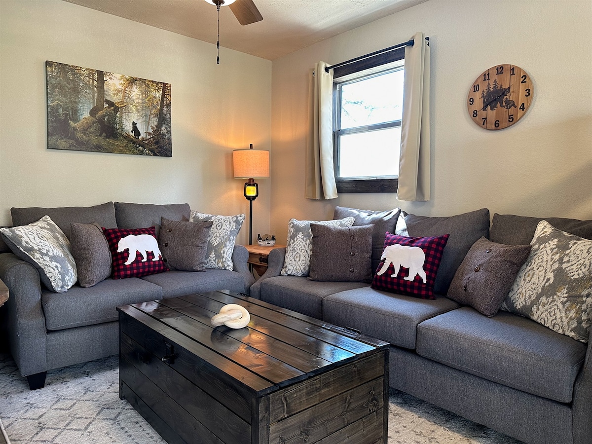 A welcoming living area features a generous gray sectional sofa adorned with patterned pillows. A rustic wooden coffee table is placed in front, and natural light filters in through a window dressed with light curtains. A nature-themed wall art and clock add a cozy touch.