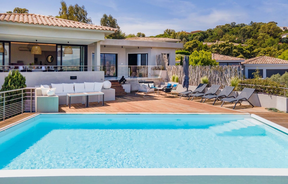 A modern villa is featured with a private pool surrounded by a spacious wooden deck. Comfortable lounge chairs are positioned around the pool, while a cozy seating area is visible near the entrance of the house. Expansive glass doors connect the indoor living space to the outdoors.