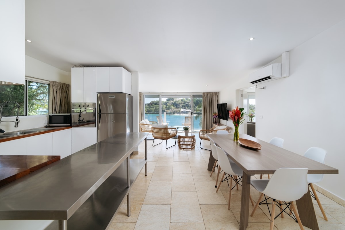 A bright and airy kitchen area features stainless steel appliances, white cabinetry, and a spacious dining table. Comfortable seating is provided by woven chairs, with large windows allowing for ample natural light and a view of the surrounding landscape.