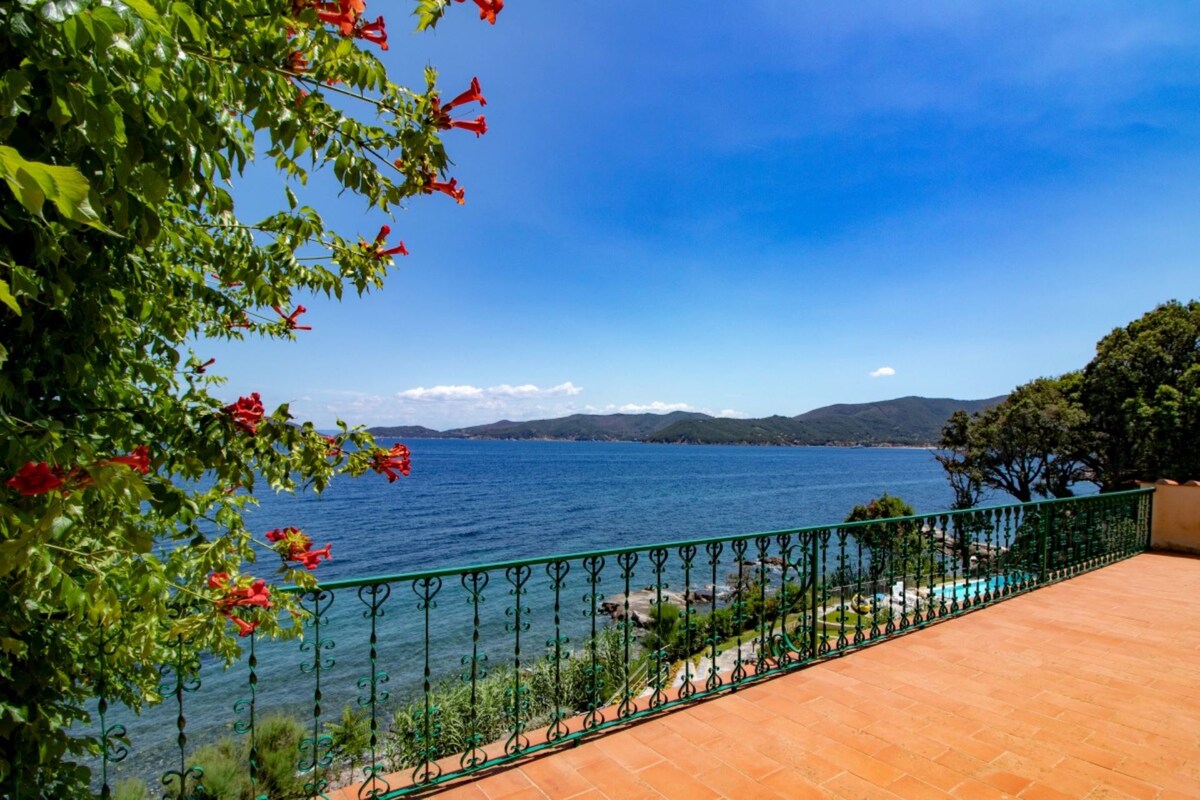 A sunlit terrace is framed by vibrant green foliage and red flowers, overlooking calm waters. The view extends across the Gulf, with mountains visible in the distance under a clear blue sky.