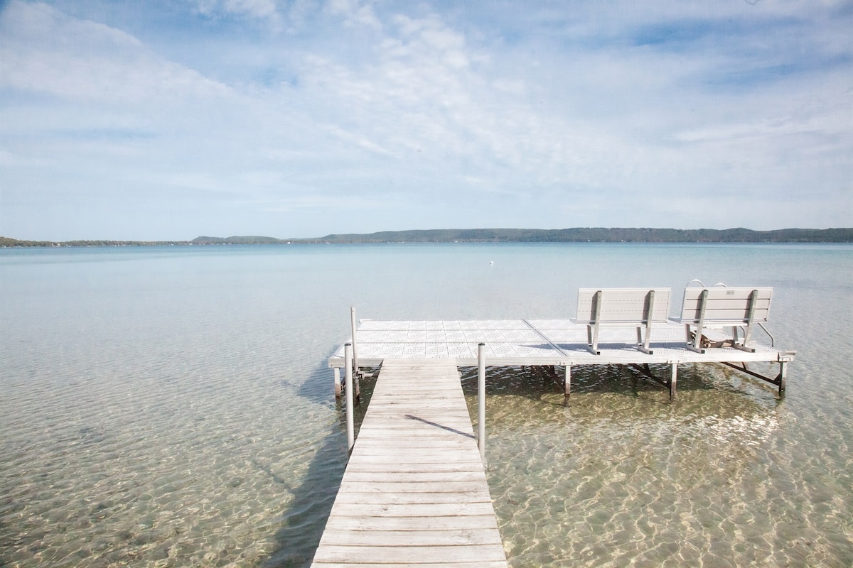 A wooden dock extends into clear waters, where gentle ripples reflect the sky. Three lounge chairs are positioned at the end of the dock, providing a serene spot for relaxation and enjoying the view of the tranquil lake and distant shoreline.