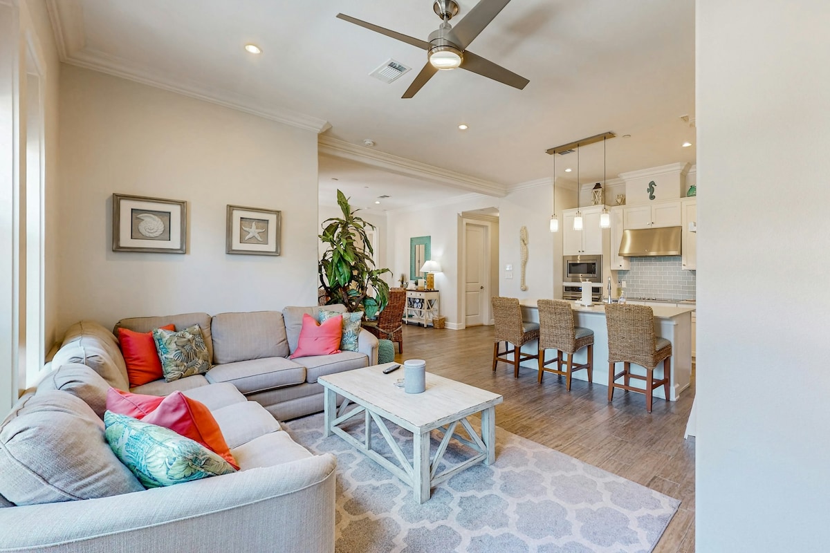 The open living area features a spacious sectional sofa adorned with bright pillows, positioned near a light-colored coffee table. A ceiling fan circulates air above, while natural light floods in through large windows. The adjoining kitchen and dining area are visible, offering a welcoming atmosphere.