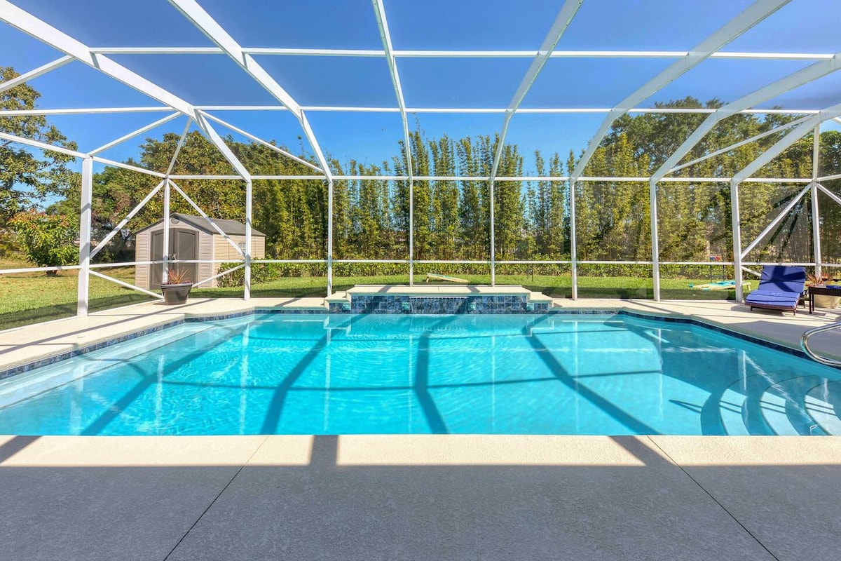 A large, heated pool is surrounded by a white screened enclosure, reflecting bright blue water under the clear sky. A waterfall feature adds a tranquil sound as sunlight casts patterns across the pool deck and surrounding grassy area.