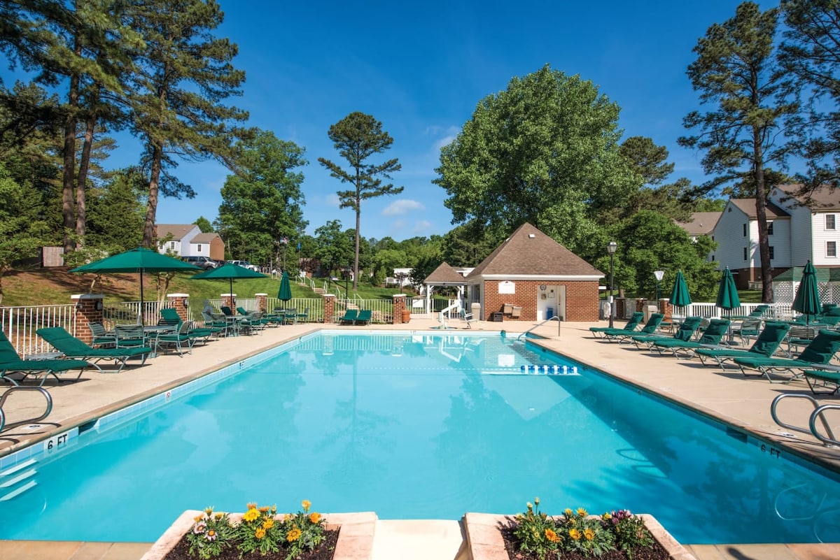 An outdoor swimming pool is framed by sun loungers and umbrellas, with vibrant flowers placed along the edge. A shaded pavilion is visible in the background, surrounded by lush greenery and residential buildings, providing a relaxing atmosphere for guests.