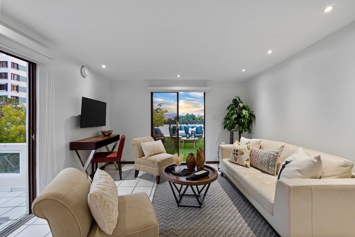 The living area features a light-colored sofa and armchair arranged around a round coffee table. A flat-screen TV is mounted on the wall, while floor-to-ceiling glass doors lead to the outdoor space, showcasing views of the surrounding greenery and sunset.