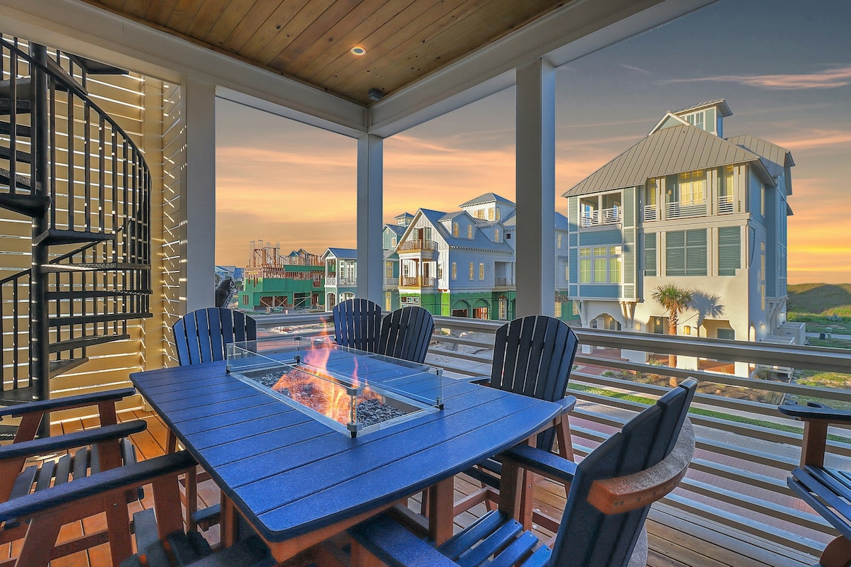 A spacious balcony is depicted, featuring a table with a central firepit surrounded by dark wooden chairs. A spiral staircase leads to an upper level, with coastal homes visible in the background against a colorful sunset.