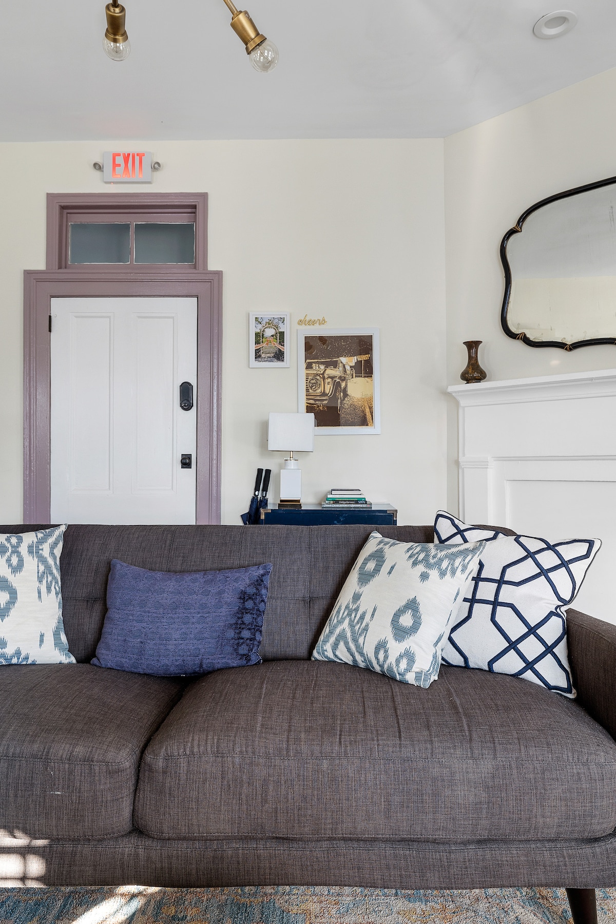 A well-appointed living area features a dark gray sofa adorned with patterned throw pillows in blue and white. A stylish mirror hangs above a decorative mantle, while a small lamp and framed artwork enhance the space. Light enters through a nearby door.