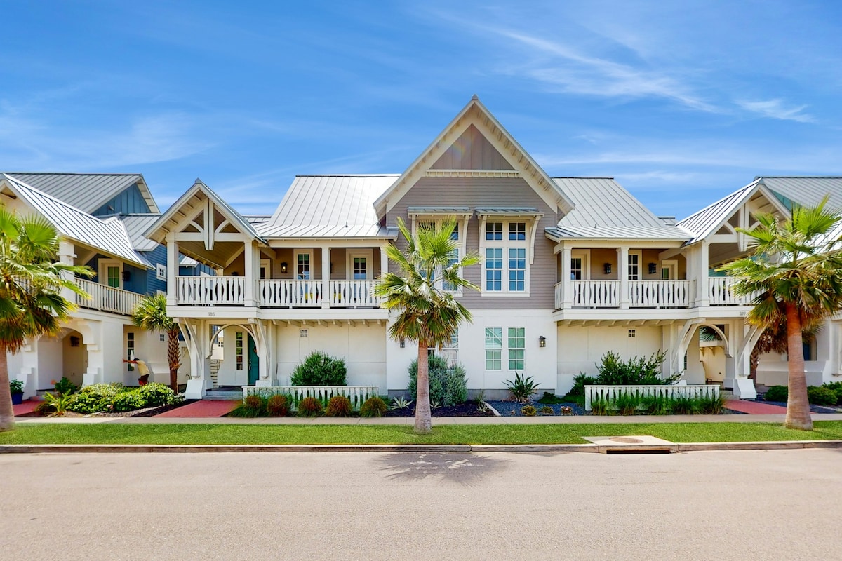 The exterior of a two-story beach home is displayed, featuring a light-colored façade with a sloped metal roof and a wide, inviting porch. Palm trees and landscaped shrubs line the walkways, adding to the coastal atmosphere. A clear blue sky completes the serene setting.