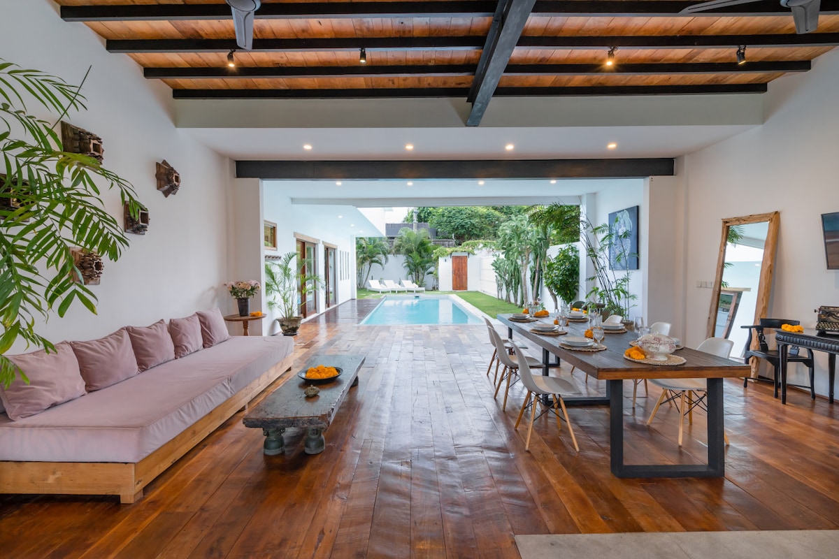 A spacious living and dining area features warm wooden flooring and exposed beams overhead. A large, comfortable sofa runs along one side, while a dining table set for meals sits across from it. Large glass doors provide a view of the lagoon-style pool and surrounding greenery.