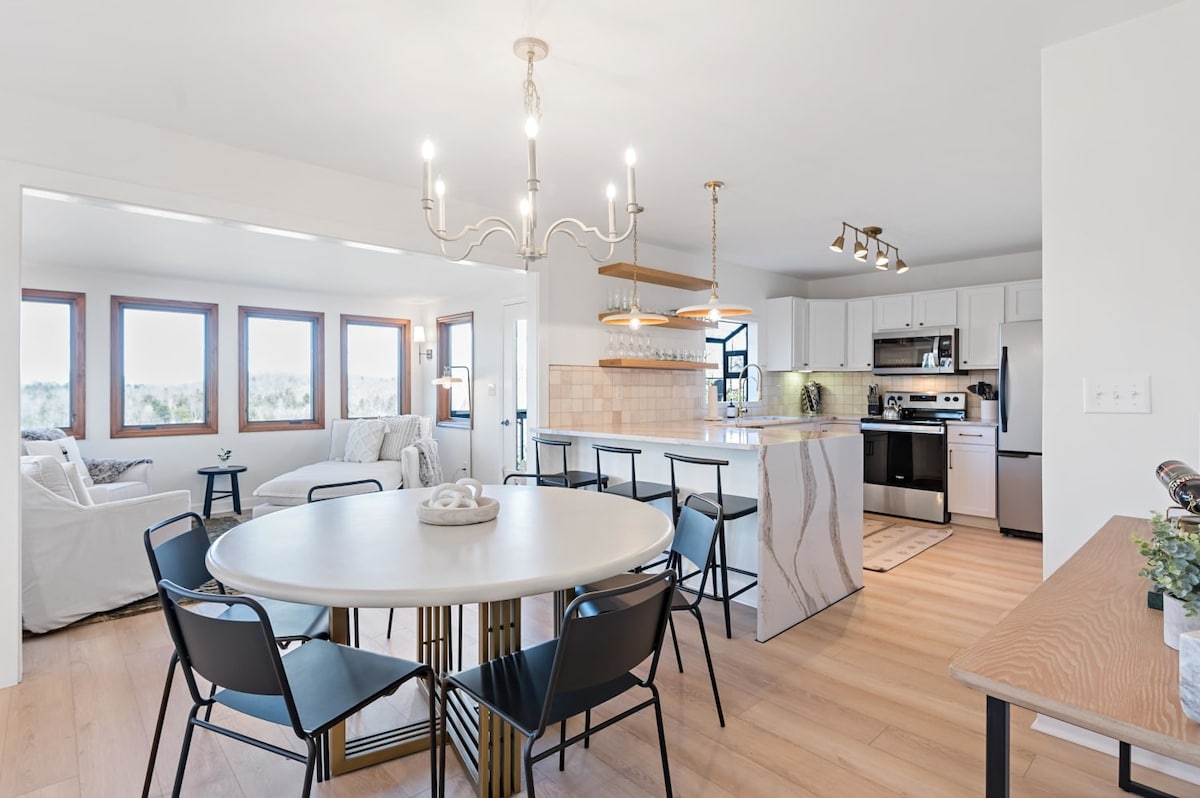 A modern open-concept kitchen and dining area feature a circular table with black chairs. Natural light floods in through large windows, illuminating the sleek countertops and stainless-steel appliances. A stylish chandelier hangs above the dining table, enhancing the inviting atmosphere.