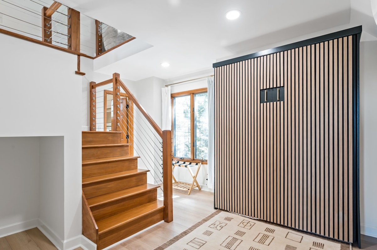 A bright entryway features polished wooden stairs leading to the upper level. The walls are adorned with vertical wooden slats, and large windows allow natural light to fill the space. A small foldable table is positioned near the window, ideal for convenience.