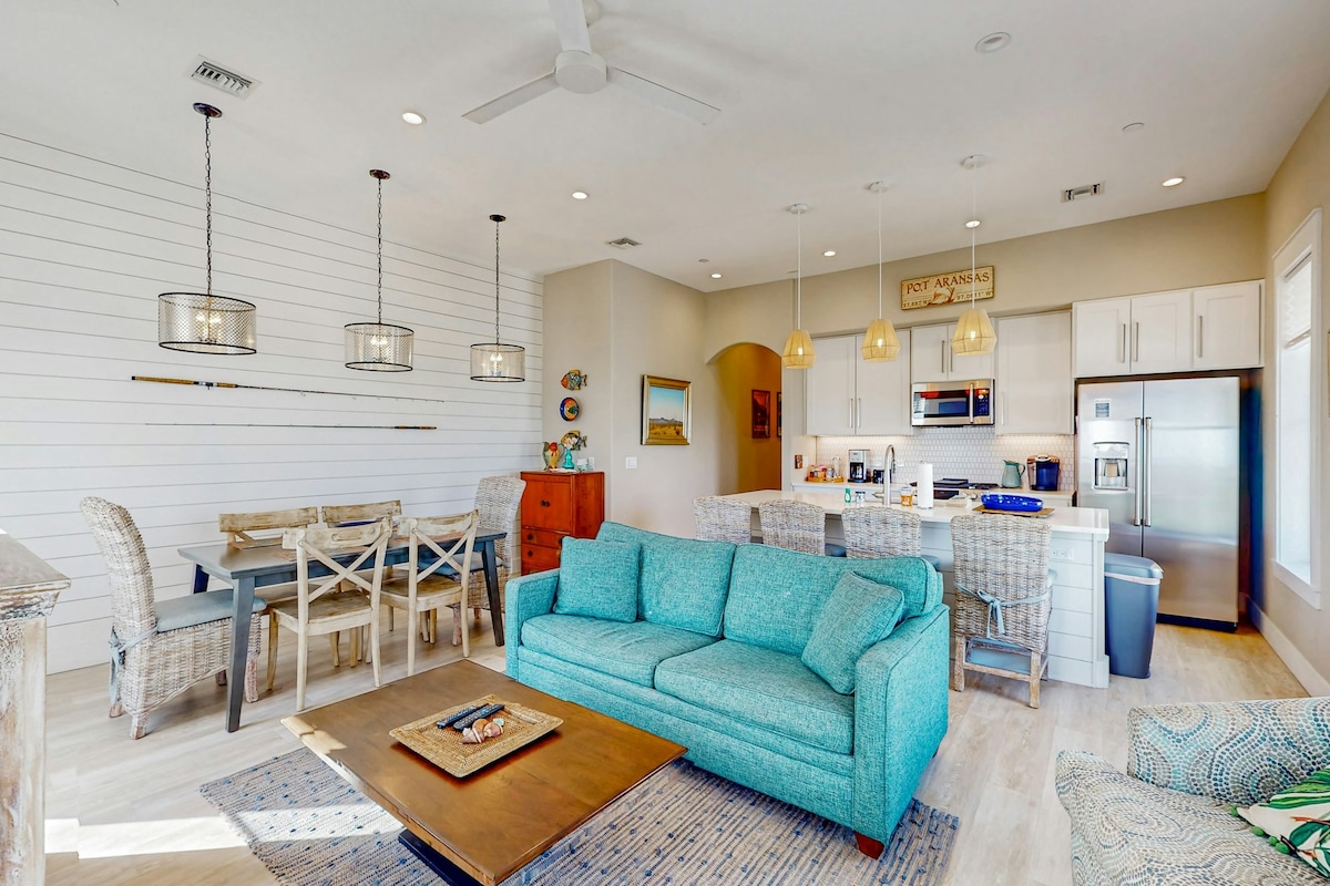 The open-concept living area features a light-filled space with a turquoise sofa, a coffee table, and a dining table set for six. Modern pendant lights hang above, and a well-equipped kitchen is visible with stainless steel appliances and a center island.