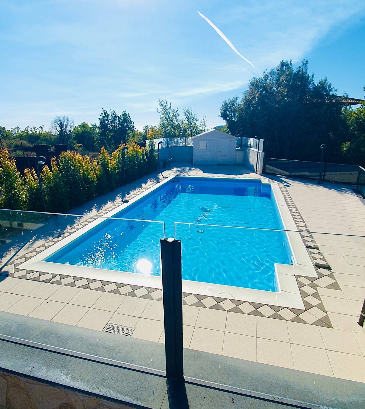 A private pool is centrally featured within a beautifully landscaped area, surrounded by greenery and trees. The pool is framed by a tiled patio, offering ample space for relaxation. A soft blue hue reflects from the clear water under bright sunlight, enhancing the outdoor ambiance.