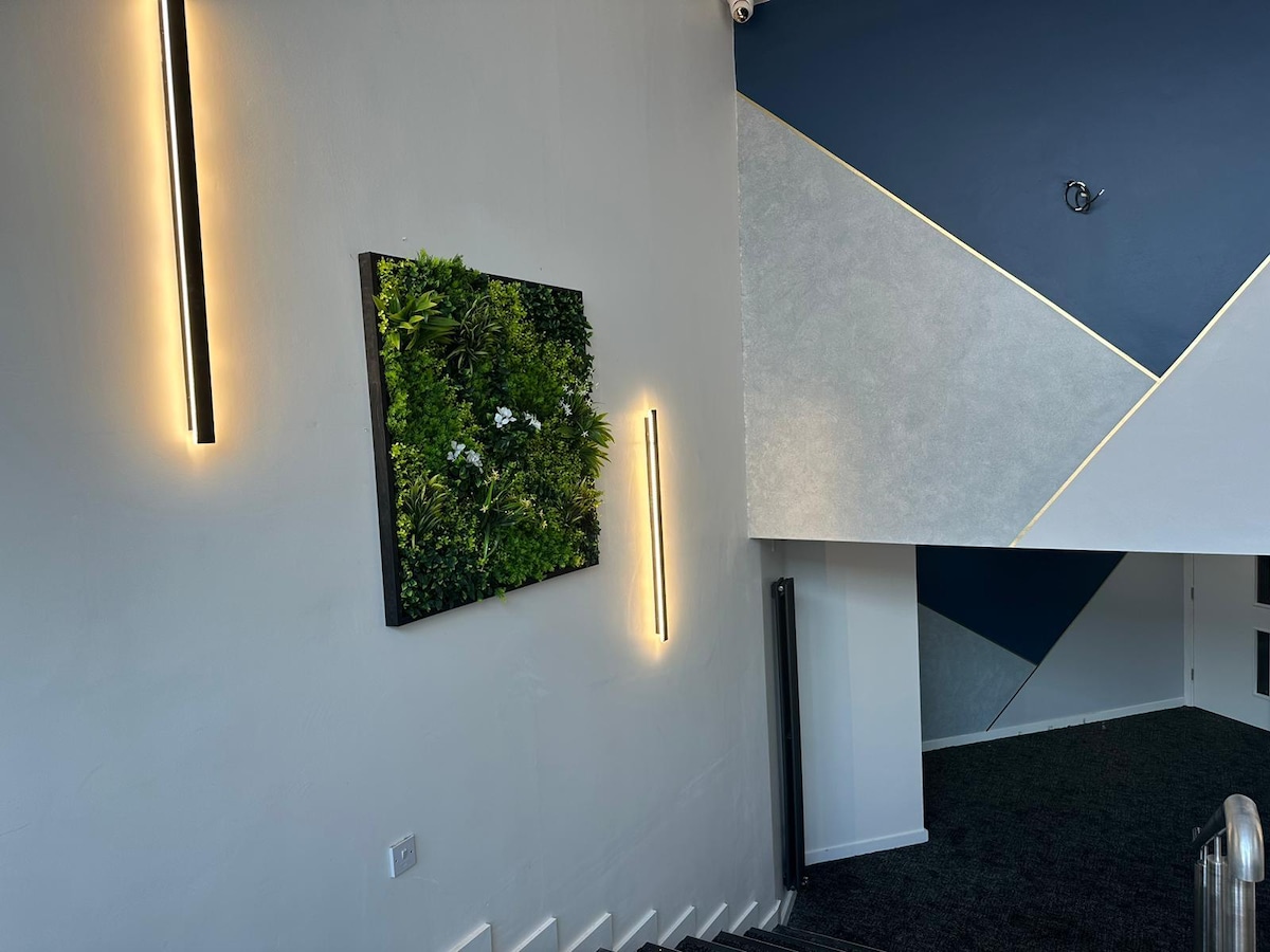 A well-lit staircase is framed by modern wall sconces. A vibrant green wall art piece composed of various plants adds a touch of nature to the space. The walls are painted in neutral tones with bold geometric accents.