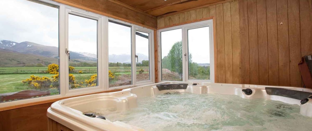 An indoor hot tub is situated beside large windows, allowing natural light and views of the surrounding landscape. The bubbling water invites relaxation, while the wooden panelling enhances the rustic charm of the space.