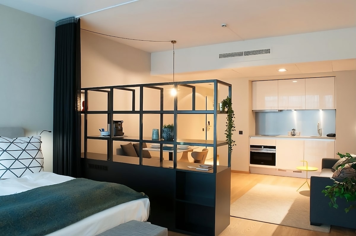 A modern studio layout is displayed, featuring a partitioned sleeping area with a spacious bed and decorative pillows. The kitchen area is well-equipped and includes a seating area, illuminated by soft lighting. Neutral tones and clean lines create a welcoming environment.
