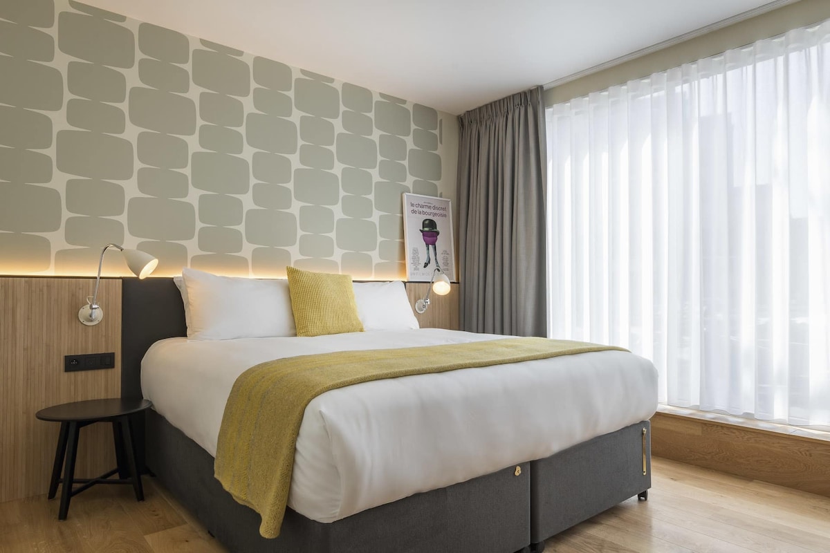 A spacious room features a king bed with a light-colored blanket and a decorative pillow. A side table stands beside the bed, and natural light filters through sheer white curtains, illuminating the patterned wall behind the bed.