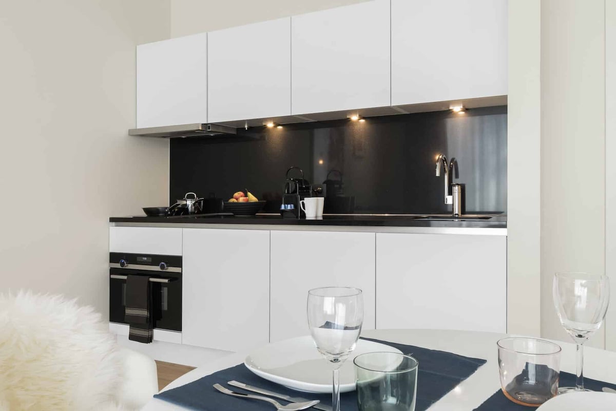 A modern kitchen area is equipped with sleek white cabinets and black countertops. A stovetop, oven, and various kitchen appliances are visible. Dining items are neatly placed on a table, complemented by glassware and a fruit bowl arranged on the counter.