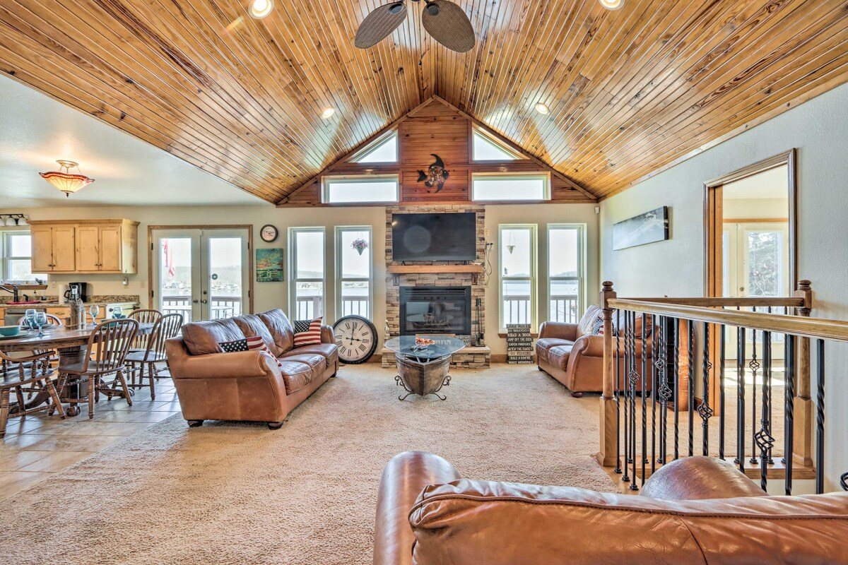 An open common area showcases a vaulted ceiling and large windows, allowing ample natural light. Comfortable leather seating is arranged around a central coffee table, with a fireplace and wall-mounted TV present. A dining area with a wooden table adds to the welcoming space.