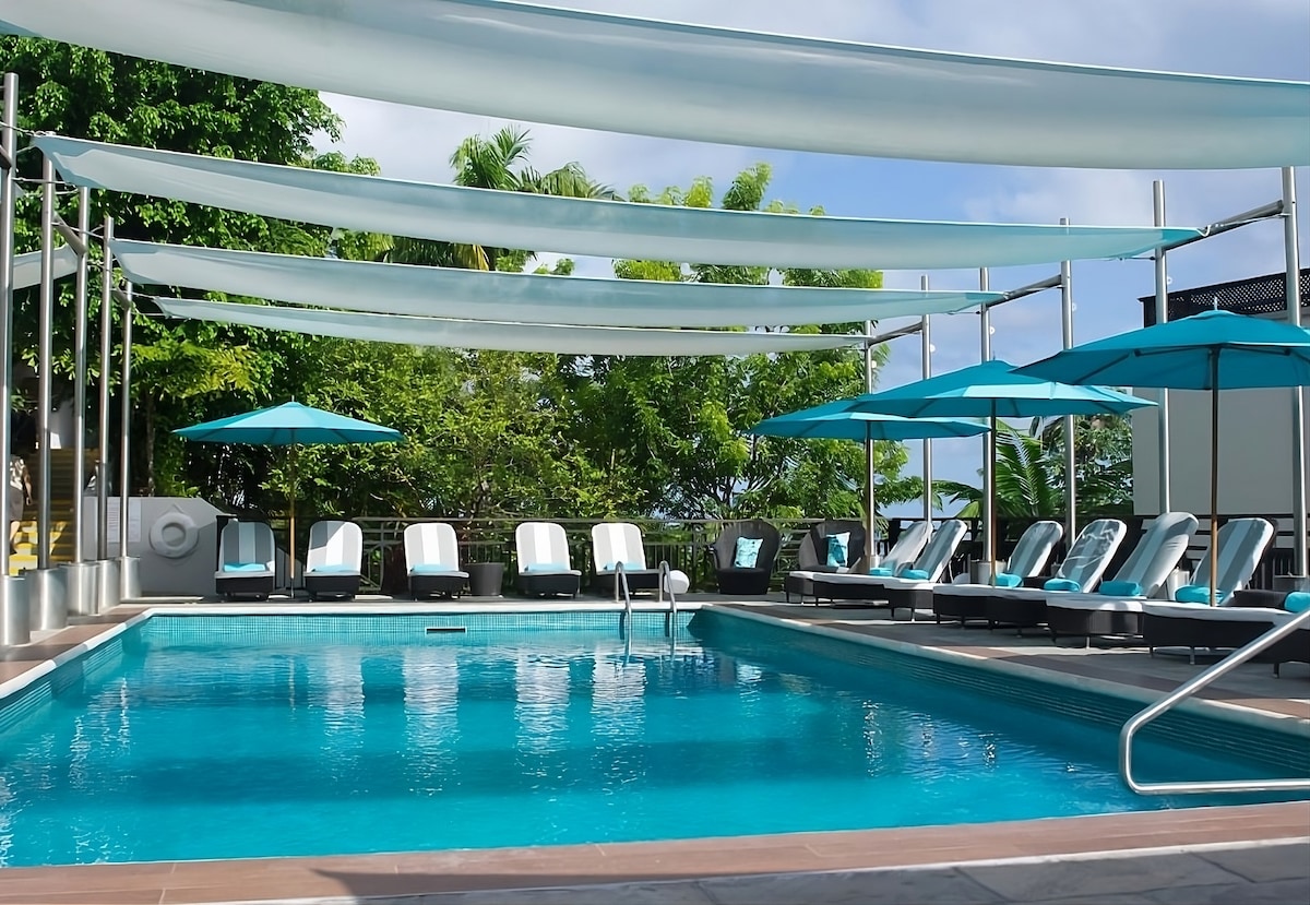 A tranquil swimming pool area features sun loungers under blue umbrellas, surrounded by greenery. The pool is shaded by fabric canopies, providing a refreshing atmosphere for relaxation. The pool's surface reflects the sky, inviting guests to unwind and enjoy the serene environment.