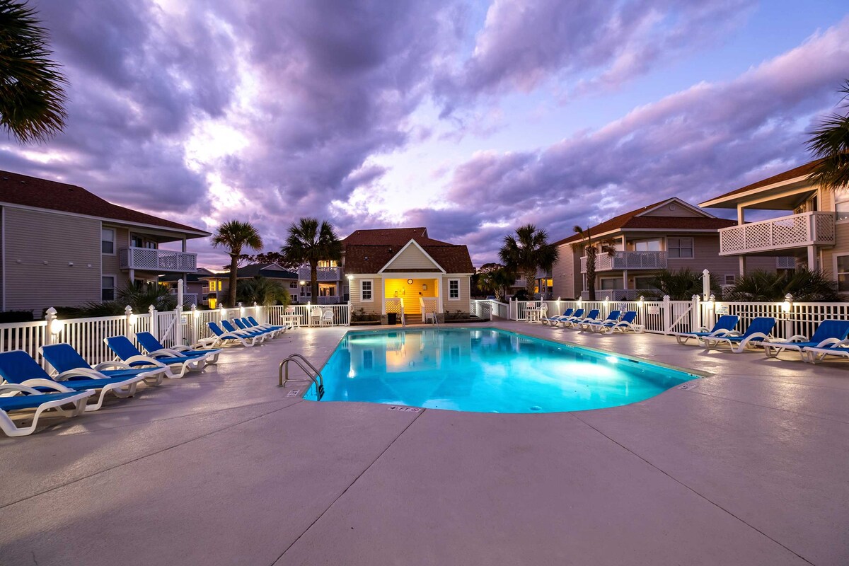 A community pool is surrounded by lounge chairs, featuring a serene blue water hue. The area is enhanced by palm trees and residential buildings, with clouds reflecting the colors of sunset above. Soft outdoor lighting creates a welcoming atmosphere.
