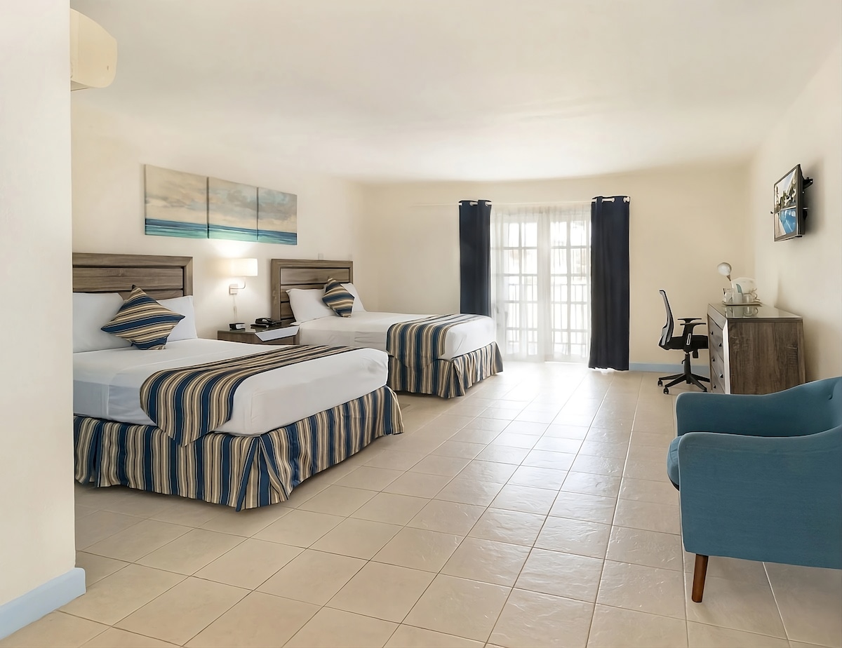 The spacious hotel room contains two double beds with striped blue and beige bedspreads. A desk with a chair and a flat-screen TV are positioned against the wall. Natural light enters through large glass doors, leading to a balcony. Neutral-colored walls complement the tile floor.
