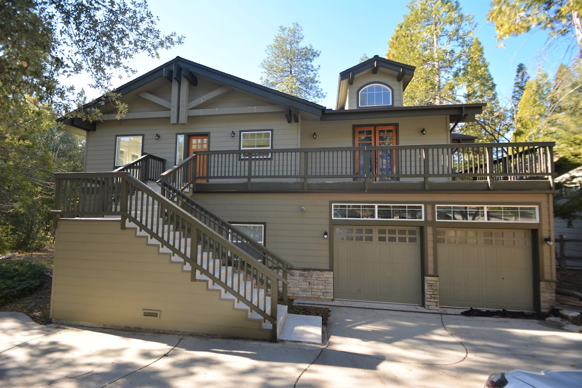 The exterior of the home is showcased, featuring a two-story design with a sloped roof. A wide staircase leads to a deck with multiple entry doors. Below, a two-car garage is visible. Surrounding greenery adds to the tranquil setting.