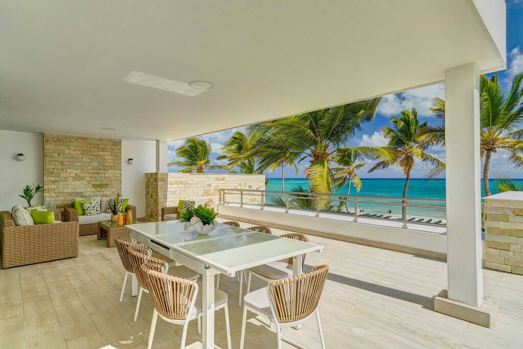 A spacious terrace features an outdoor dining table surrounded by stylish chairs. Comfortable seating is positioned nearby, with lush greenery and vibrant ocean views beyond. Sun-drenched palm trees sway gently against a bright sky, enhancing the tranquil coastal atmosphere.