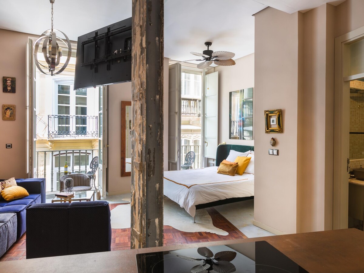 A light-filled living space is showcased with a spacious bed and a blue sofa, complemented by a decorative ceiling fan. Large windows open to the charming street view, while a television is mounted on the partitioned wall, enhancing the room's functionality.