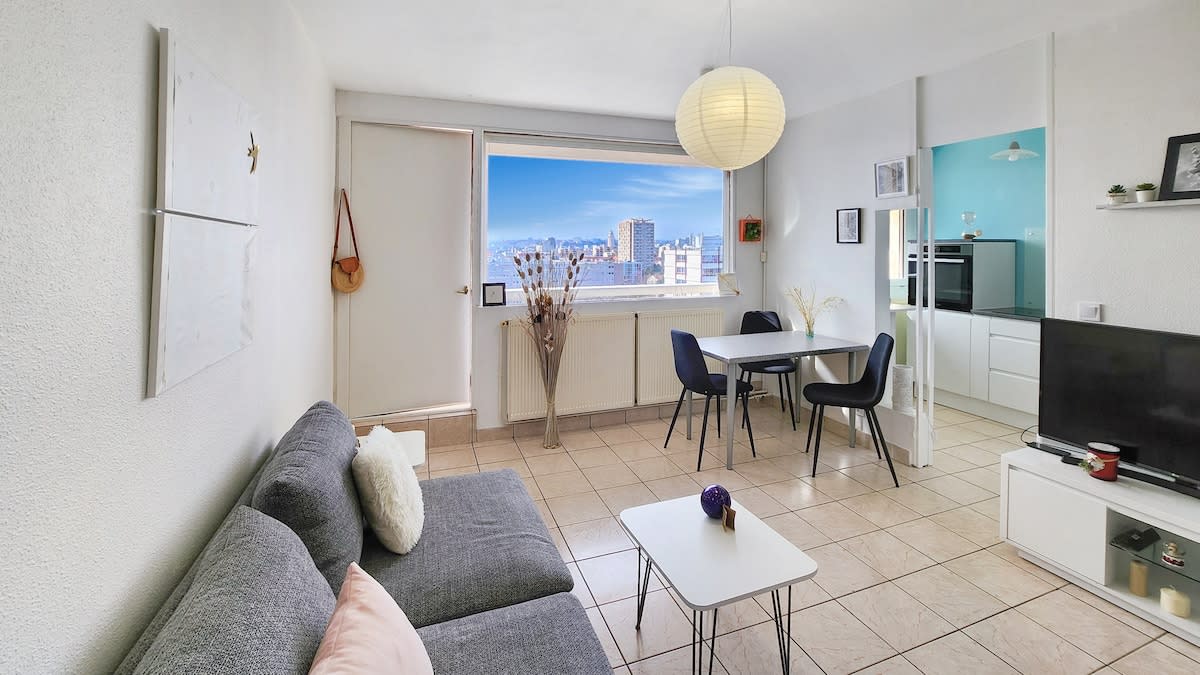 A functional living area features a spacious gray sofa facing a television. A round dining table accommodates four, positioned near a large window with panoramic city views. Neutral decor and natural light create an inviting atmosphere, complemented by a modern pendant light.