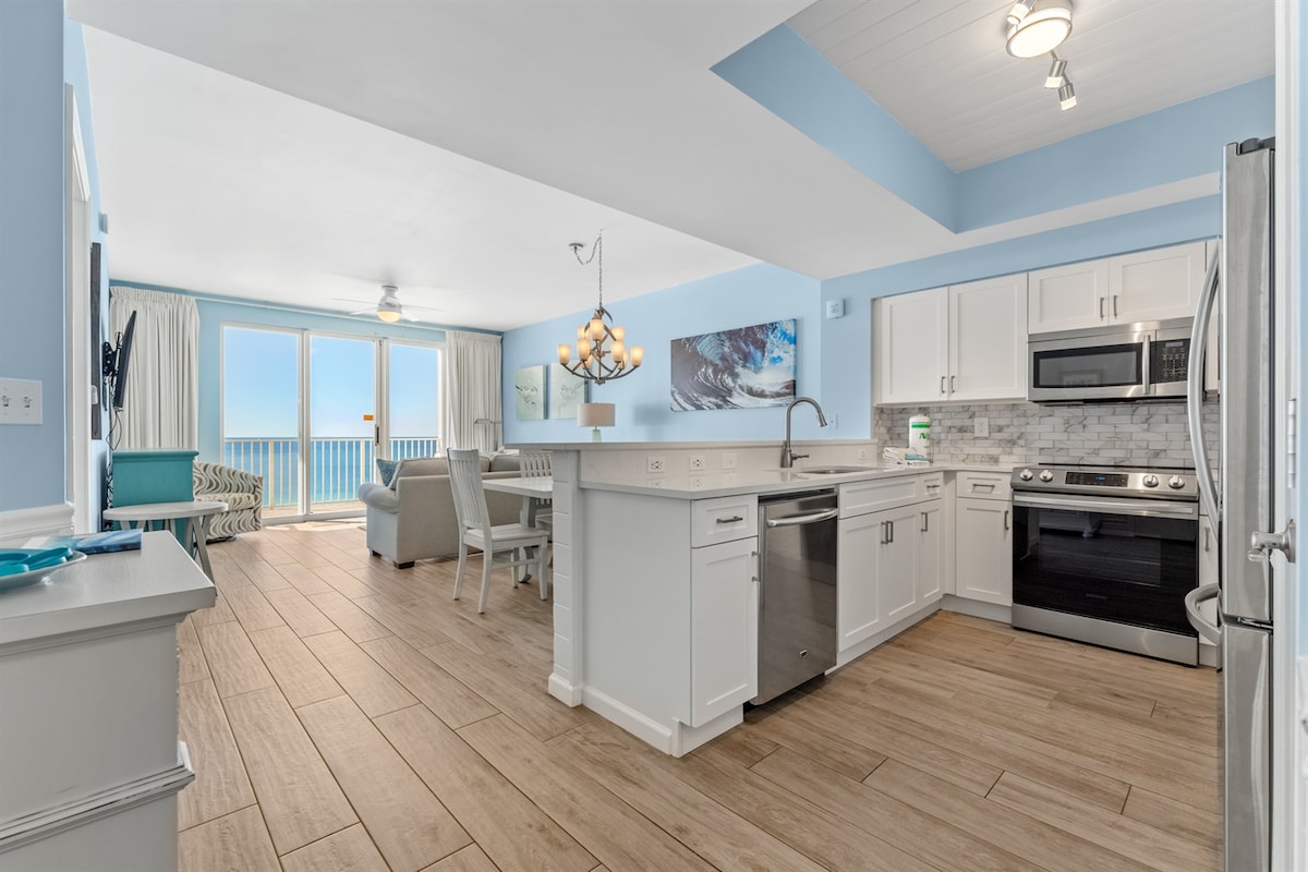 An updated kitchen and living area are depicted, featuring light cabinetry and a spacious island. Large sliding glass doors provide views of the Gulf, allowing natural light to illuminate the space. Comfortable seating arrangements are visible, framed by a soft blue wall.