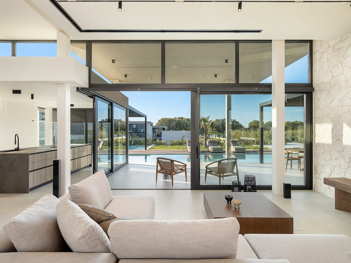 A modern living space displays large glass doors that open to a serene outdoor area. Natural light fills the interior, highlighting a comfortable seating area with a soft sofa and coffee table. The view includes a swimming pool and lush greenery in the background.
