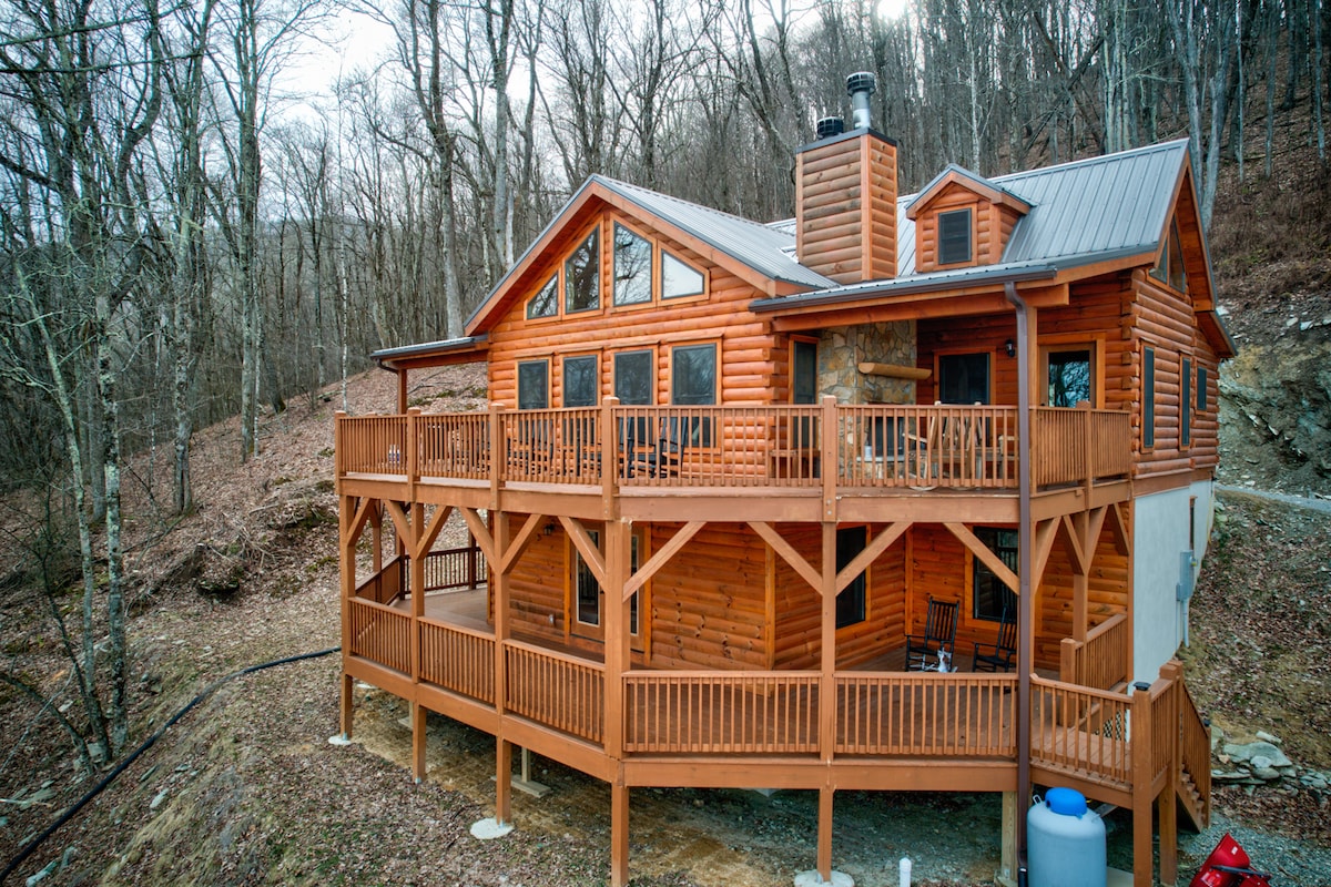The spacious log cabin is framed by surrounding trees, presenting a welcoming wooden exterior with multiple large windows. A broad deck wraps around the structure, equipped with wooden railings and seating areas, providing an inviting space to enjoy the natural surroundings.