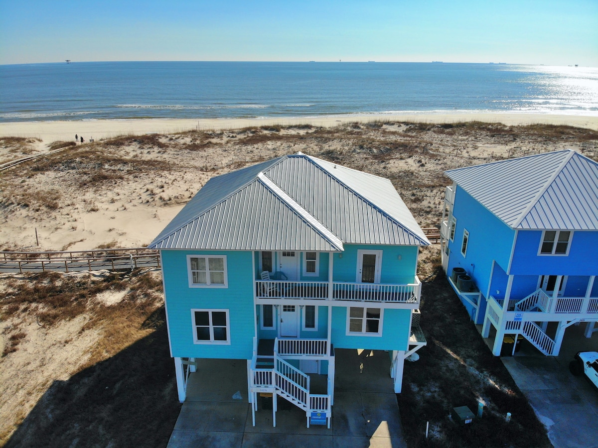A two-story beachfront home is showcased in a vibrant blue color, standing adjacent to the sandy shore. The elevated structure includes expansive balconies, with staircases leading to the main entrance. The serene ocean and clear sky outline the property, emphasizing its prime seaside location.
