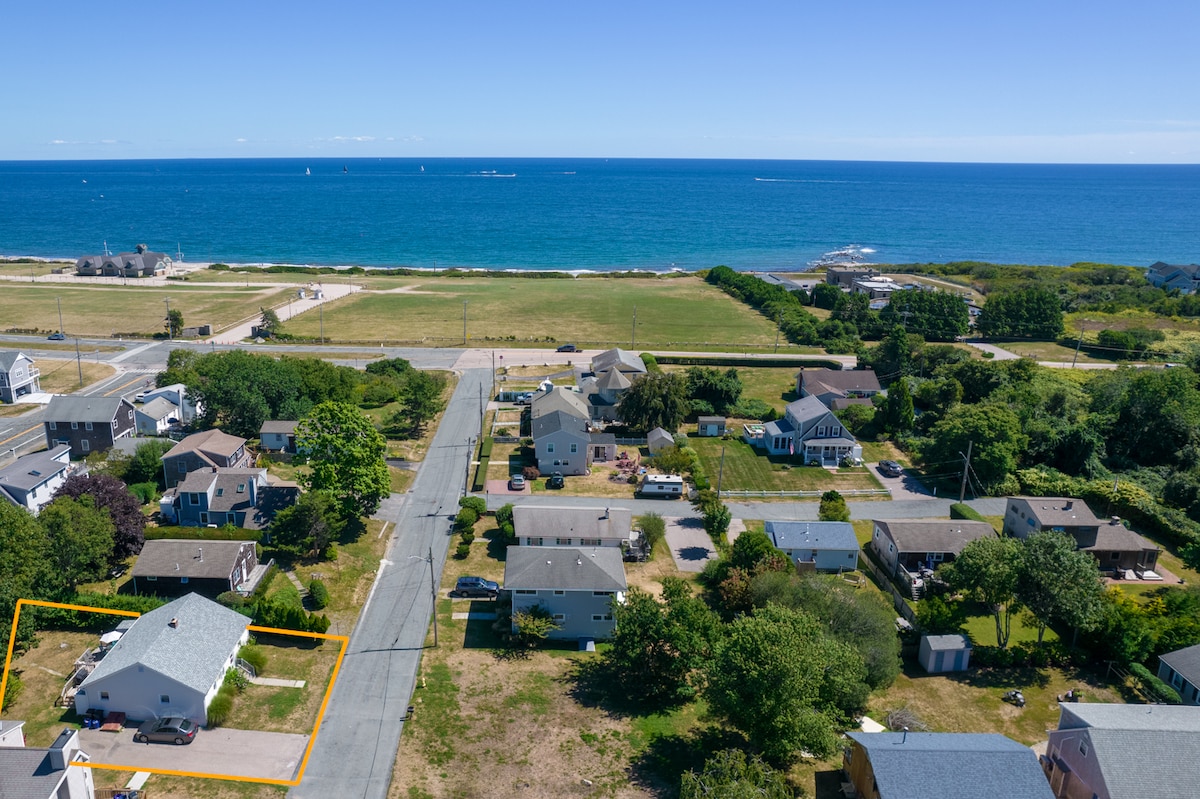 An aerial view shows the vacation home in a quiet neighborhood, with a clear view of the ocean in the background. Multiple homes and green spaces are visible, along with a road that leads to the beach. The immediate area appears spacious and accessible.