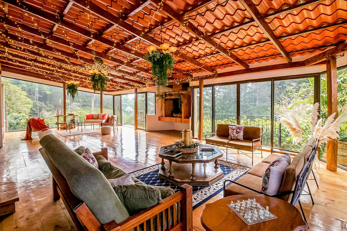The spacious living room features a wooden ceiling adorned with string lights, enhancing the bright ambiance. Floor-to-ceiling windows provide expansive views of the surrounding jungle, while comfortable seating arrangements invite relaxation. A central wooden coffee table holds a chess set, contributing to the inviting atmosphere.