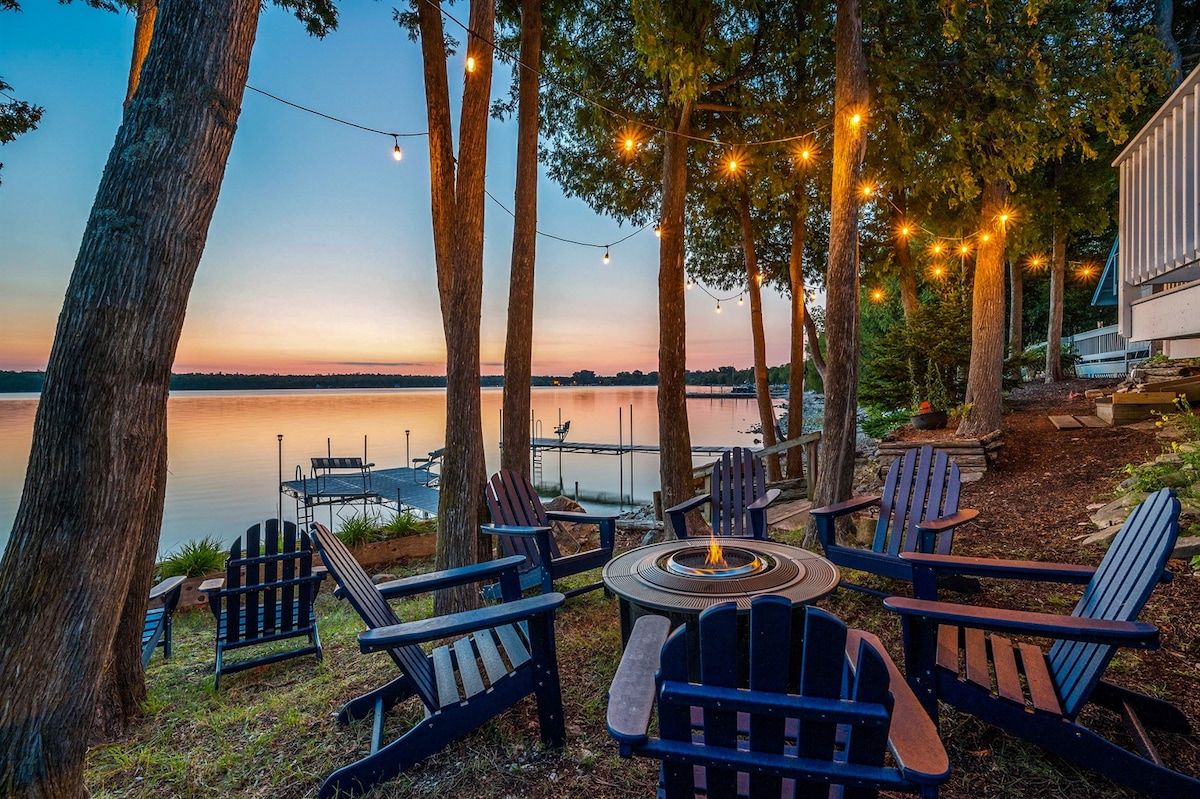 An outdoor fire pit area is surrounded by several wooden adirondack chairs. Soft lights are strung between trees, casting a warm glow. The serene bay and dock are visible in the background, reflecting colors of the sunset.