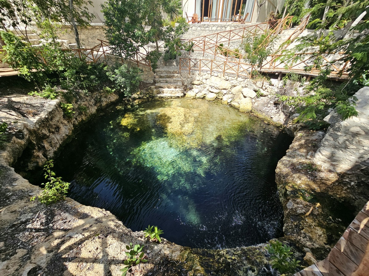 A natural water feature is surrounded by an earthy stone edge and lush greenery. The water exhibits various shades of blue and emerald, reflecting the sunlight. A wooden pathway can be seen leading to the water's edge, enhancing the serene environment.