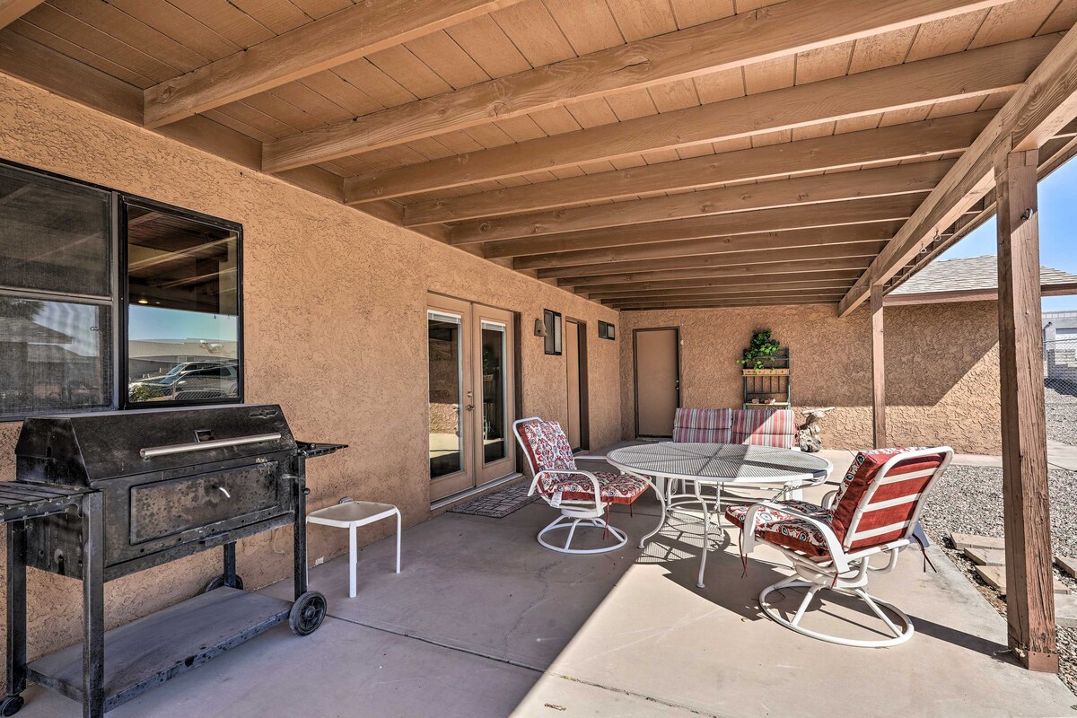 An outdoor patio area features a circular dining table surrounded by several chairs, including one with a cushion. A grill is positioned nearby under a wooden overhang, while the space is framed by large windows allowing natural light. Decorative elements are visible on the walls.
