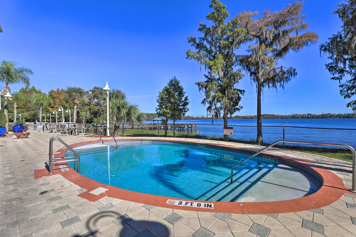A refreshing outdoor pool is surrounded by a paved patio, featuring comfortable lounge chairs. Lush greenery and cypress trees create a natural backdrop, while a serene lake view enhances the ambiance. Clear blue skies provide a bright, inviting atmosphere.