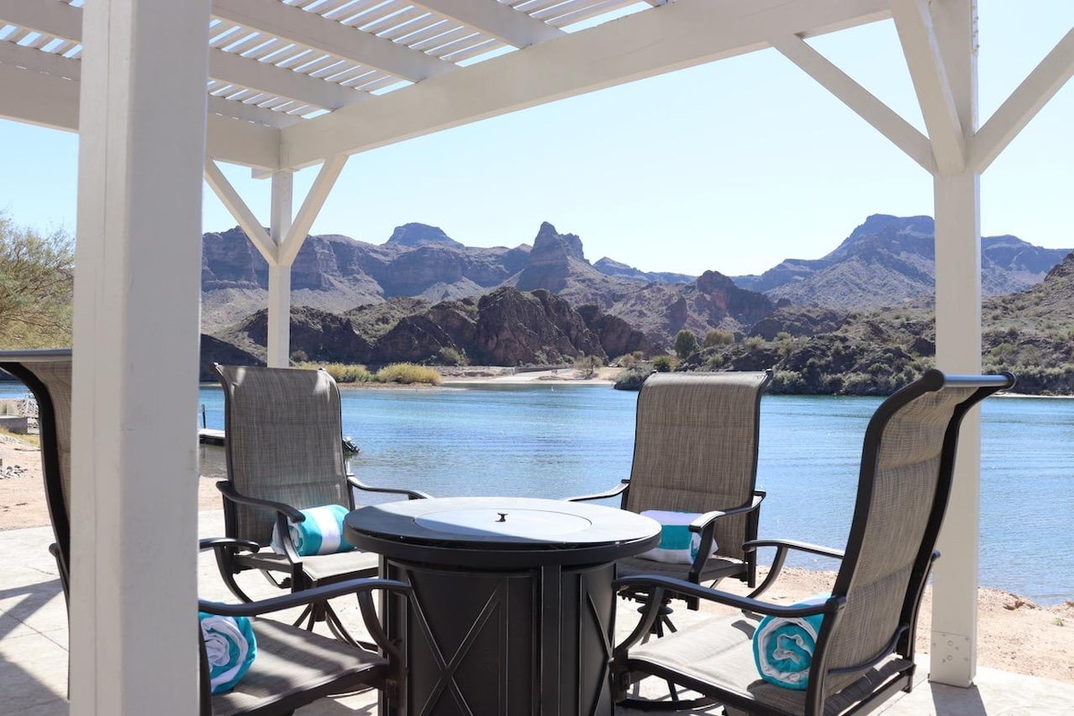A shaded outdoor seating area features four comfortable chairs arranged around a circular fire pit table. The scenic backdrop showcases the calm river and surrounding mountains, offering a serene view that invites relaxation.