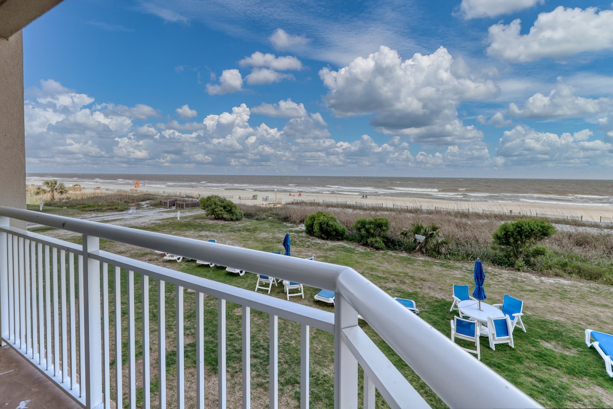 The private balcony offers unobstructed ocean views and features several blue lounge chairs. The grassy area below is dotted with additional seating, with the beach visible in the distance. A clear sky with white clouds frames the picturesque coastal scene.
