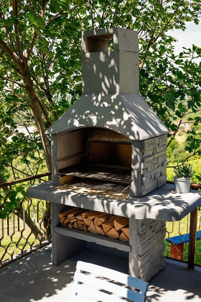 A stone barbecue is positioned on a shaded terrace, surrounded by greenery. The structure features a grill area above with a chimney, while a lower shelf holds neatly stacked firewood. Sunlight filters through the leaves, casting playful shadows on the barbecue.