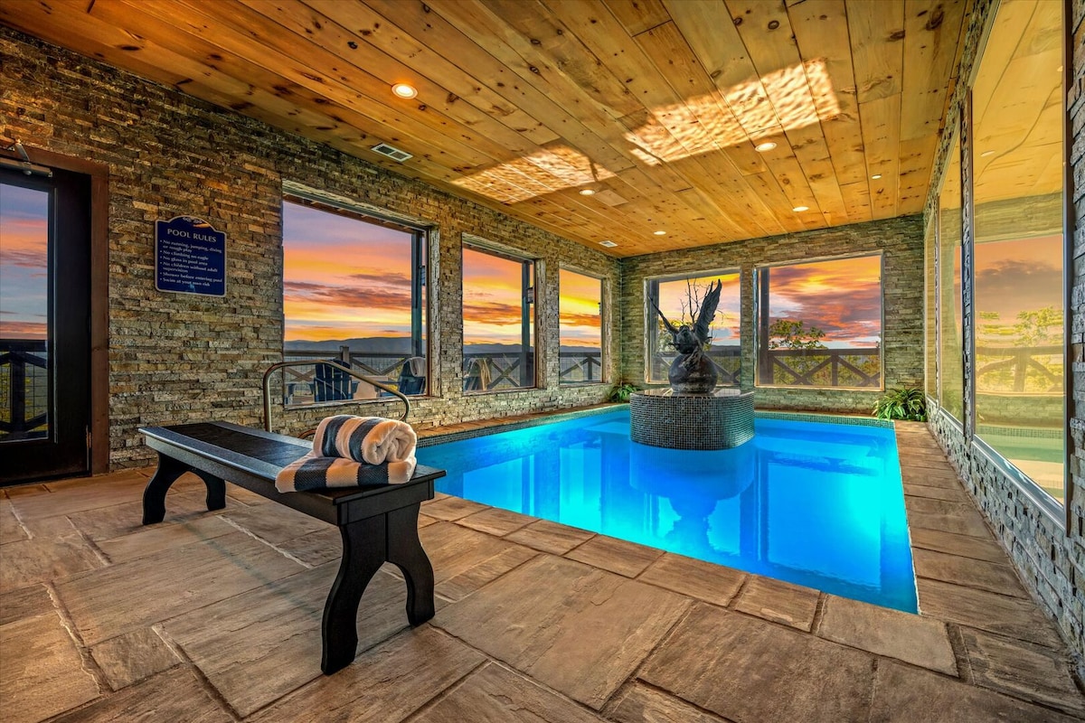 An indoor saltwater pool is highlighted under a wooden ceiling, with stone walls framing expansive windows showcasing a vibrant sunset. A stone fountain featuring an eagle sculpture complements the tranquil blue water, providing a serene atmosphere for relaxation.
