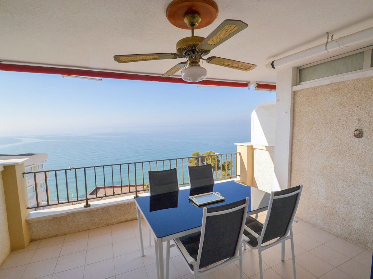 A terrace features a dining table with six chairs, overlooking a calm sea. A ceiling fan is installed above, and the space is bordered by protective railings. The view showcases a serene landscape, enhancing the outdoor experience.