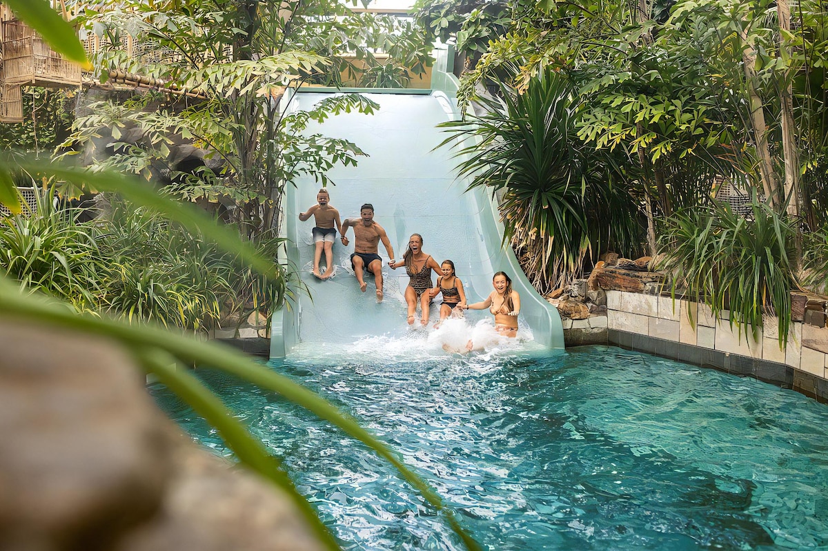 A water slide surrounded by lush greenery offers a fun experience, as several individuals descend into a sparkling pool below. Tropical plants frame the scene, creating a natural oasis that enhances the playful atmosphere of the water feature.