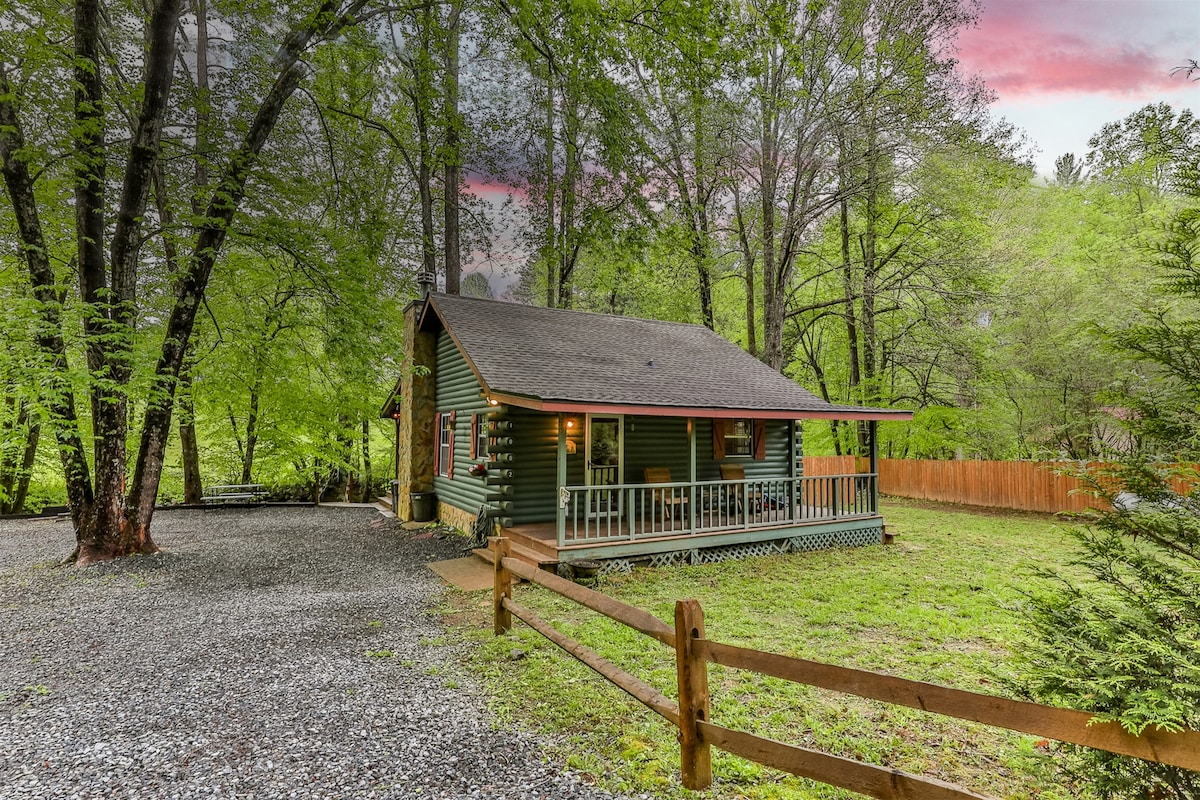 A rustic cabin is set among tall trees, featuring a green exterior and a welcoming porch. A gravel path leads up to the cabin, surrounded by a grassy area and a wooden fence. Scenic views of nature are visible in the background.